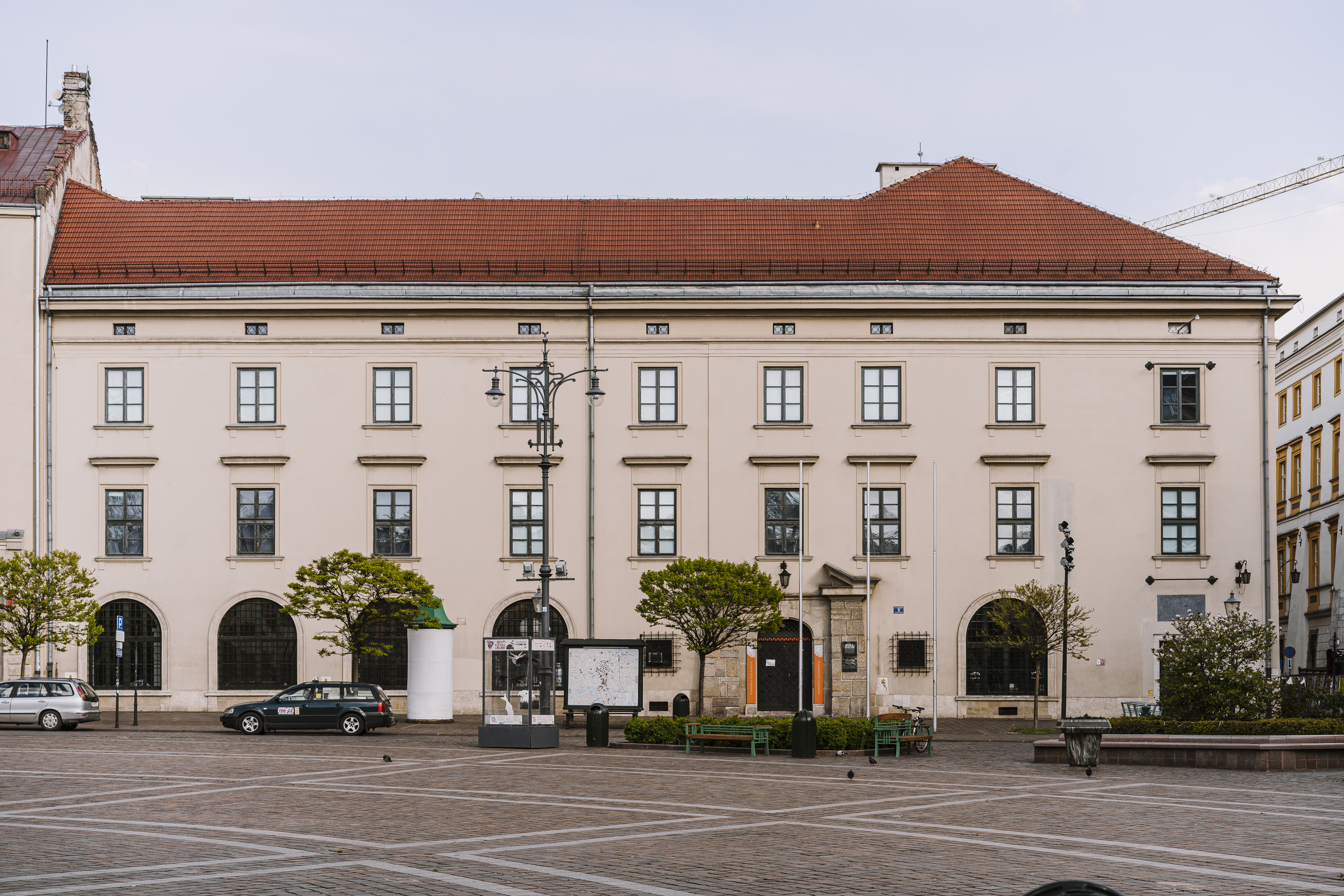 Duży jasnobeżowy trzykondygnacyjny budynek z czerwonym dachem i symetrycznymi oknami oraz łukowymi witrynami na parterze, frontem na szeroki brukowany plac z latarnią uliczną, kioskiem z mapą, trzema małymi drzewkami, ławkami oraz kilkoma zaparkowanymi samochodami.