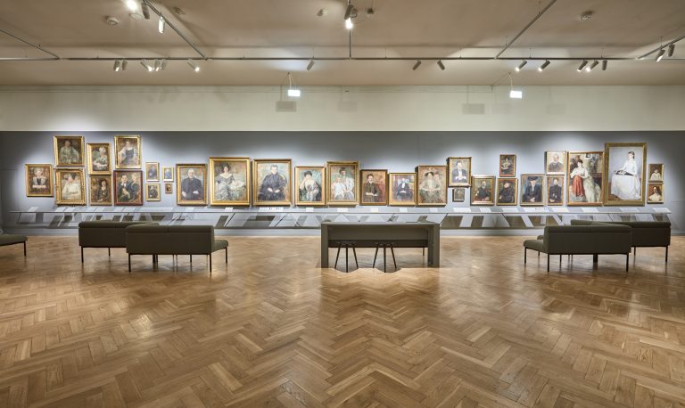 Duża galeria muzealna z długą szarą ścianą pokrytą licznymi portretami w zlotych ramach ułożonymi w rzędzie, pod nimi niskie półki z podpisami, na pierwszym planie kilka szarych ławek i stół z krzesłami, drewniana jodełkowa podłoga i sufitowe reflektory oświetlające obrazy.
