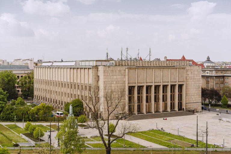 Panoramiczne zdjęcie miejskie pokazujące duży prostokątny kamienny budynek z wysokimi pionowymi filarami i szerokimi schodami prowadzącymi na główny plac, na dachu którego widoczne są maszty antenowe i szklane świetliki; przed budynkiem rozciąga się brukowany plac z kilkoma osobami i skuterem, po bokach zielone tarasy z alejkami, drzewami i pomnikiem, a w tle niskie budynki z czerwonymi dachami pod zachmurzonym niebem.