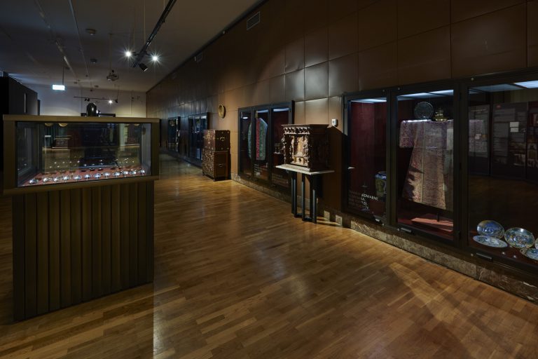 Przyciemniona sala muzealna z drewnianą podłogą i długą ścianą po prawej z przeszklonymi gablotami prezentującymi haftowaną szatę, ceramiczne talerze i rzeźbiony drewniany mebel, oraz wolnostojącymi oszklonymi gablotami po lewej i w centrum z ułożonymi łyżkami i punktowym oświetleniem sufitowym.
