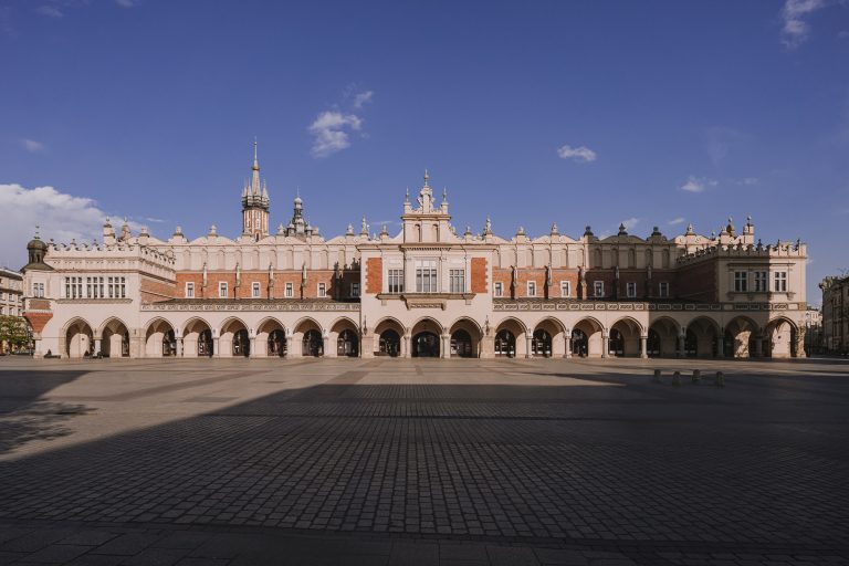 Panoramiczne przedstawienie Sukiennic na krakowskim Rynku Głównym: długa ceglasto‑jasna fasada z rzędami łukowych arkad, ozdobną attyką i wieżyczkami w tle, przed rozległym brukowanym placem pod czystym niebem.