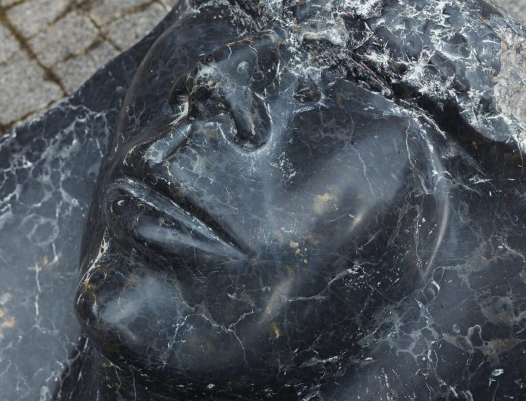 Zbliżenie na czarne, białymi żyłkami żyłkowane marmurowe oblicze rzeźby leżącej — widoczny profil nosa, zamknięte oko i usta oraz fragment kostki brukowej w tle.