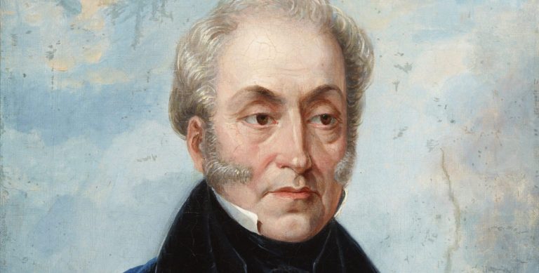 Malarski portret półpostać mężczyzny o jasnych, siwiejących włosach i bokobrodach, zwróconego trzy czwarte, ubrany w białą koszulę, czarną muszkę i ciemny płaszcz z granatowym okryciem, na jasnoniebieskim, lekko chmurowym tle.
