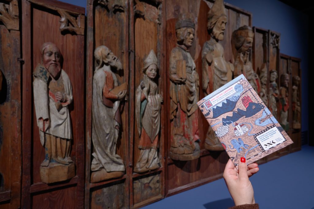 Na pierwszym planie po prawej dłoń z czerwonymi paznokciami trzyma ukośnie kolorową ulotkę z mapowo-ilustracyjnym wzorem, w tle na niebieskiej ścianie rozciąga się duży, wpionowo podzielony, zużyty drewniany panel z rzeźbionymi postaciami w długich szatach w niszach.