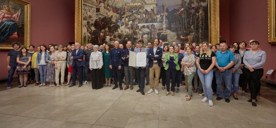 Duża grupa dorosłych ustawionych w szerokim rzędzie w sali muzealnej, na środku jedna osoba trzyma otwarty dokument, w tle duży obraz przedstawiający scenę z wieloma postaciami w złotej ramie na czerwonej ścianie, a wszyscy stoją na jasnej marmurowej podłodze w różnorodnych strojach.