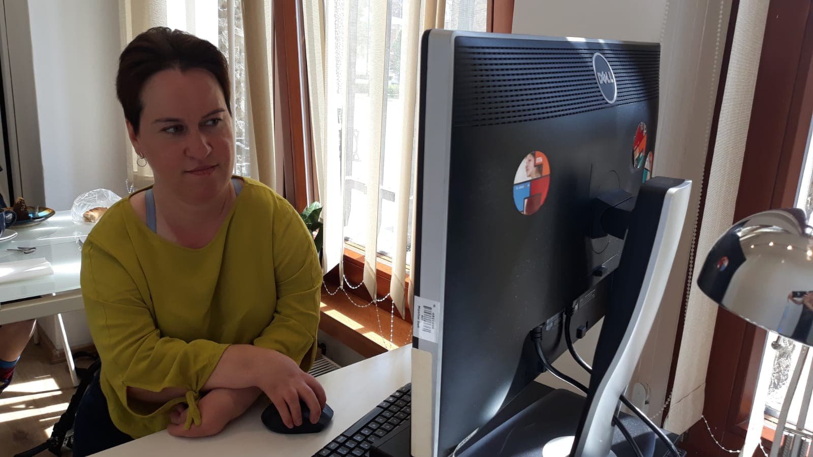 Kobieta z krótkimi ciemnymi włosami w jasnozielonej bluzce siedzi przy białym biurku po lewej i trzyma mysz komputerową, na pierwszym planie po prawej stoi duży monitor Dell, za nią widać okno z pionowymi żaluzjami, lampę biurkową i talerz z pieczywem na drugim biurku w tle.