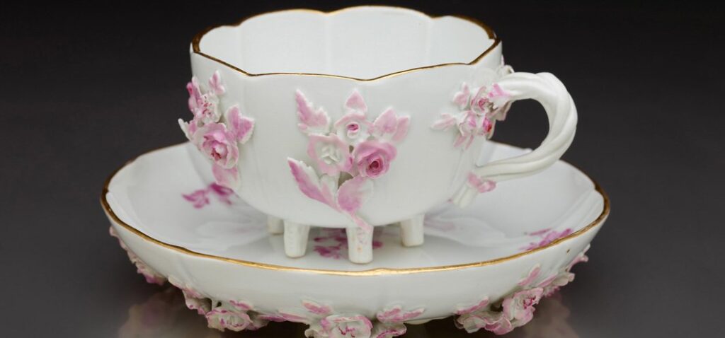 Biała porcelanowa filiżanka na trzech krótkich nóżkach ustawiona na dopasowanym spodku, obie części zdobione wypukłymi różowymi kwiatkami i cienką złotą obwódką, z ozdobnym uchem, na ciemnym tle.