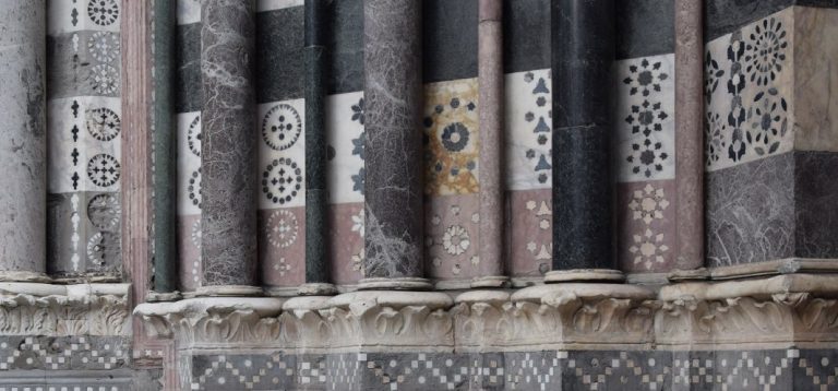 Fragment elewacji z szeregiem smukłych marmurowych kolumn w różnych odcieniach (czarny, szary, różowy) osadzonych na rzeźbionych kapitelach, za którymi widoczne są kwadratowe pola inkrustowane geometrycznymi i rozetkowymi wzorami z wielobarwnego marmuru oraz dolny fryz z drobnoziarnistą mozaiką w układzie szachownicy.
