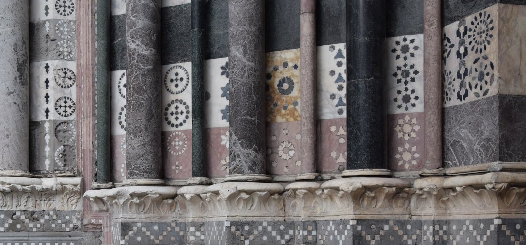 Fragment elewacji z szeregiem smukłych marmurowych kolumn w różnych odcieniach (czarny, szary, różowy) osadzonych na rzeźbionych kapitelach, za którymi widoczne są kwadratowe pola inkrustowane geometrycznymi i rozetkowymi wzorami z wielobarwnego marmuru oraz dolny fryz z drobnoziarnistą mozaiką w układzie szachownicy.