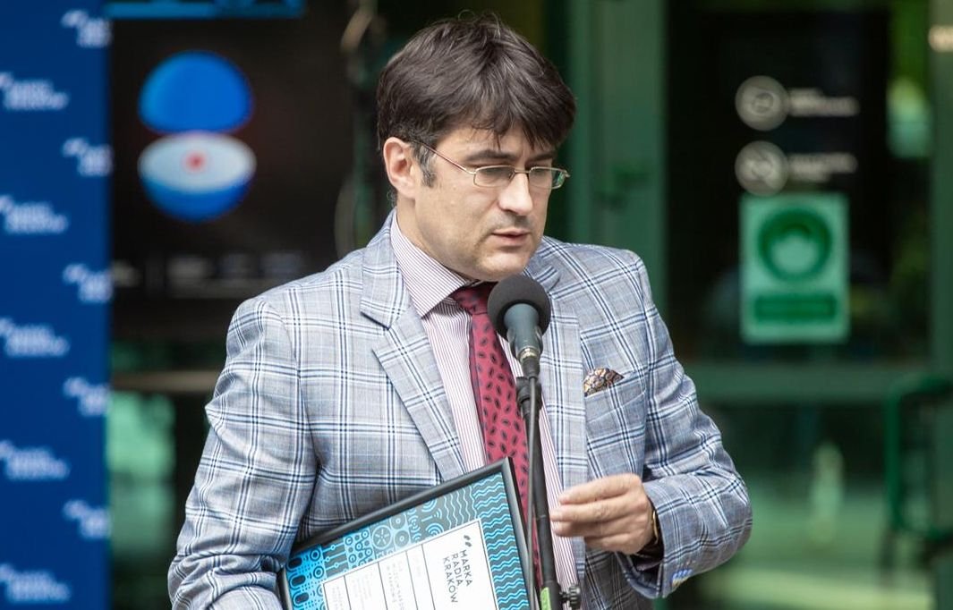 Mężczyzna w okularach w popielatej marynarce w kratę, w koszuli w cienkie paski i czerwonym krawacie stoi przy mikrofonie trzymając oprawiony dokument z niebieską grafiką, na tle przeszklonego wejścia i tablic informacyjnych.