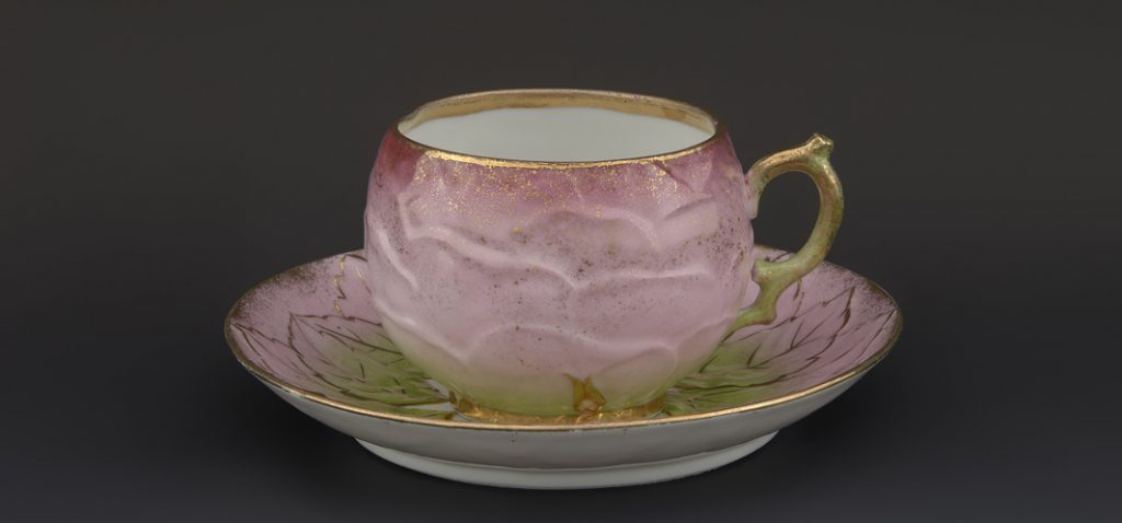 Różowa porcelanowa filiżanka o wypukłej, przypominającej płatki formie z zielonymi akcentami i złotym rantem oraz uchwytem, ustawiona na dopasowanym spodku z rysunkiem liści na ciemnym tle.