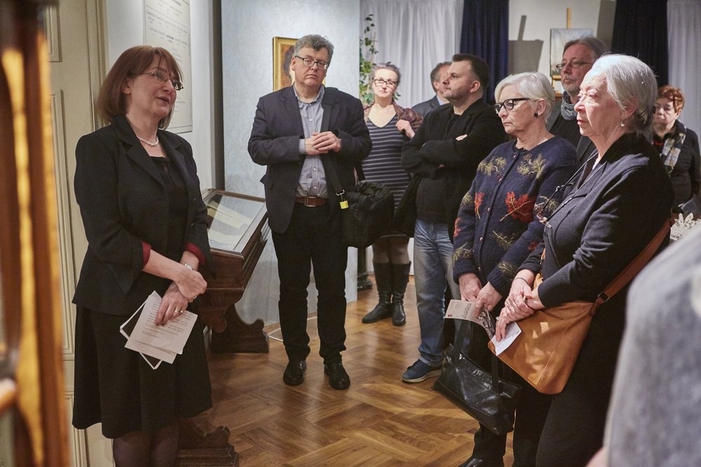 Grupa dorosłych osób stojących w wystawowym pomieszczeniu; po lewej kobieta w czarnej marynarce trzyma kartki, po prawej kilka osób w okularach i kurtkach, w tle drewniana podłoga, gabloty z eksponatami i obrazy na ścianach.