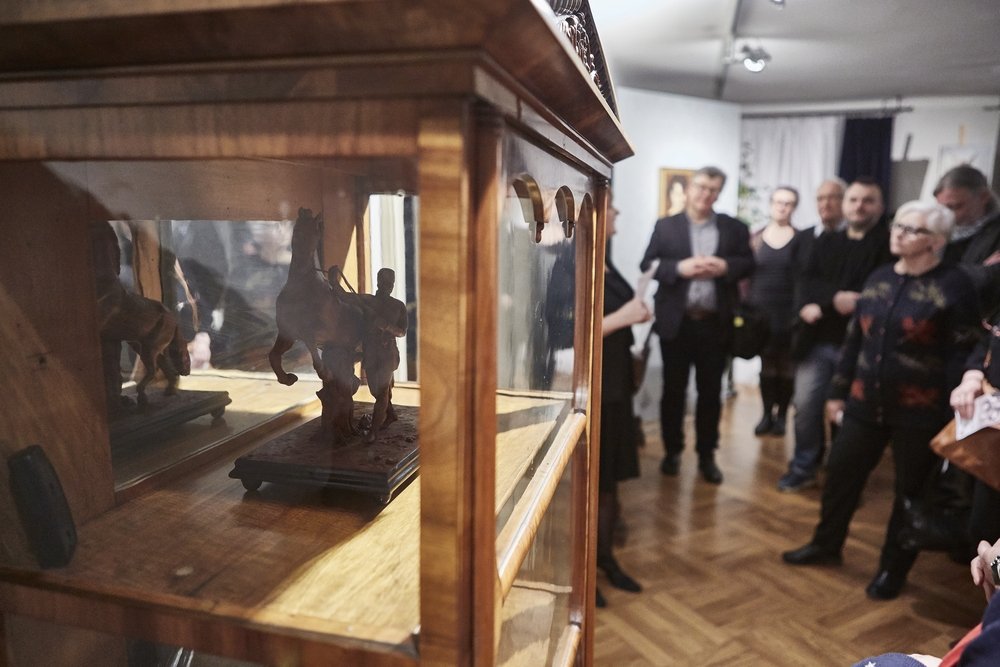 Na pierwszym planie przeszklona drewniana gablotka z małą figurką konia i postacią na podstawie odbitą w lustrze wewnątrz, w tle rozmyta grupa osób stojących na jasnej, drewnianej podłodze w oświetlonym pomieszczeniu z obrazami na ścianach.