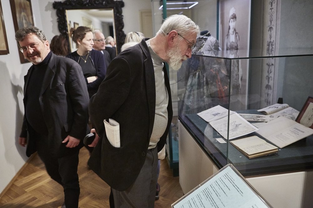 Grupa zwiedzających w muzeum; starszy mężczyzna z siwą brodą i okularami pochylony nad szklaną gablotą z otwartymi książkami i rysunkami, za nim inni odwiedzający, duże ozdobne lustro oraz wystawiona fotografia kobiety i historyczna sukienka.