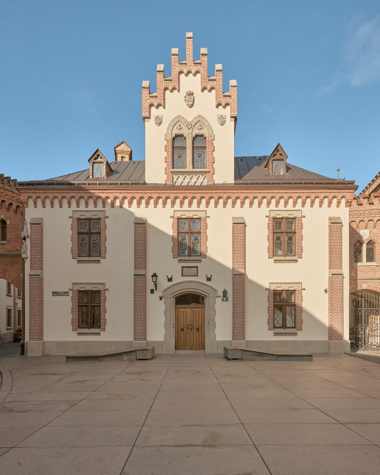 Fasada budynku o jasnobeżowej elewacji z czerwonymi ceglastymi obramowaniami, centralnym wysokim szczytem z krenelażem i kamiennymi ornamentami oraz rzeźbioną tarczą, centralnym drewnianym wejściem w łukowym portalu, symetrycznymi oknami z kratowanymi szybami, dachem z lukarnami, płaskim kamiennym placem na pierwszym planie i niskimi kamiennymi ławami pod bezchmurnym błękitnym niebem.