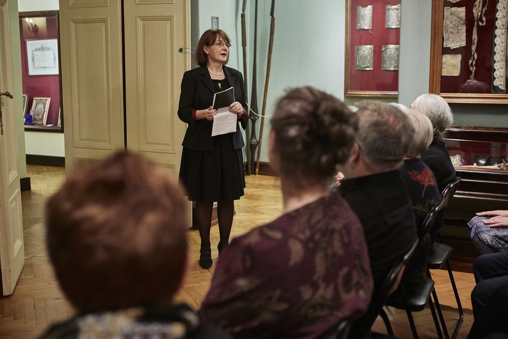 Kobieta w czarnym stroju stoi na parkietowej podłodze w sali z gablotami i ramami na ścianach, trzyma dokumenty i przemawia przed widownią widoczną z tyłu, siedzącą na krzesłach.
