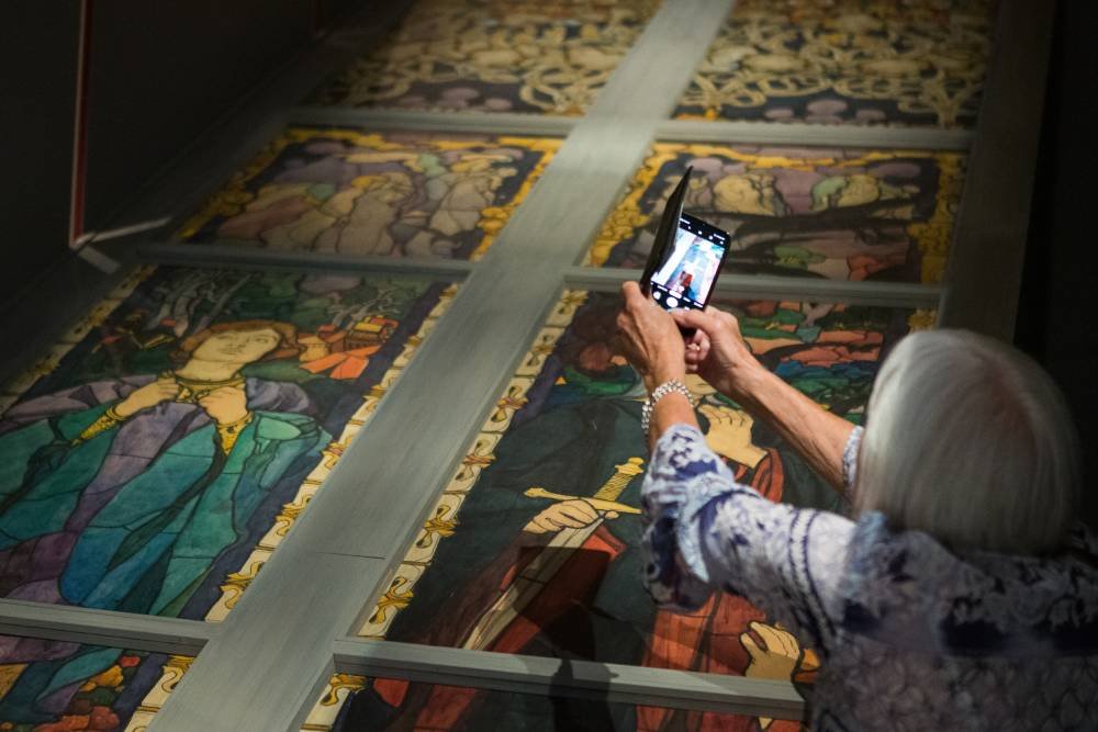 Osoba z siwymi włosami widziana od tyłu w wzorzystej bluzce i z bransoletką trzyma smartfon, fotografując kolorowe, historycznie stylizowane panele witrażowe ułożone poziomo za metalowymi ramami, na ekranie telefonu widać podgląd zdjęcia.