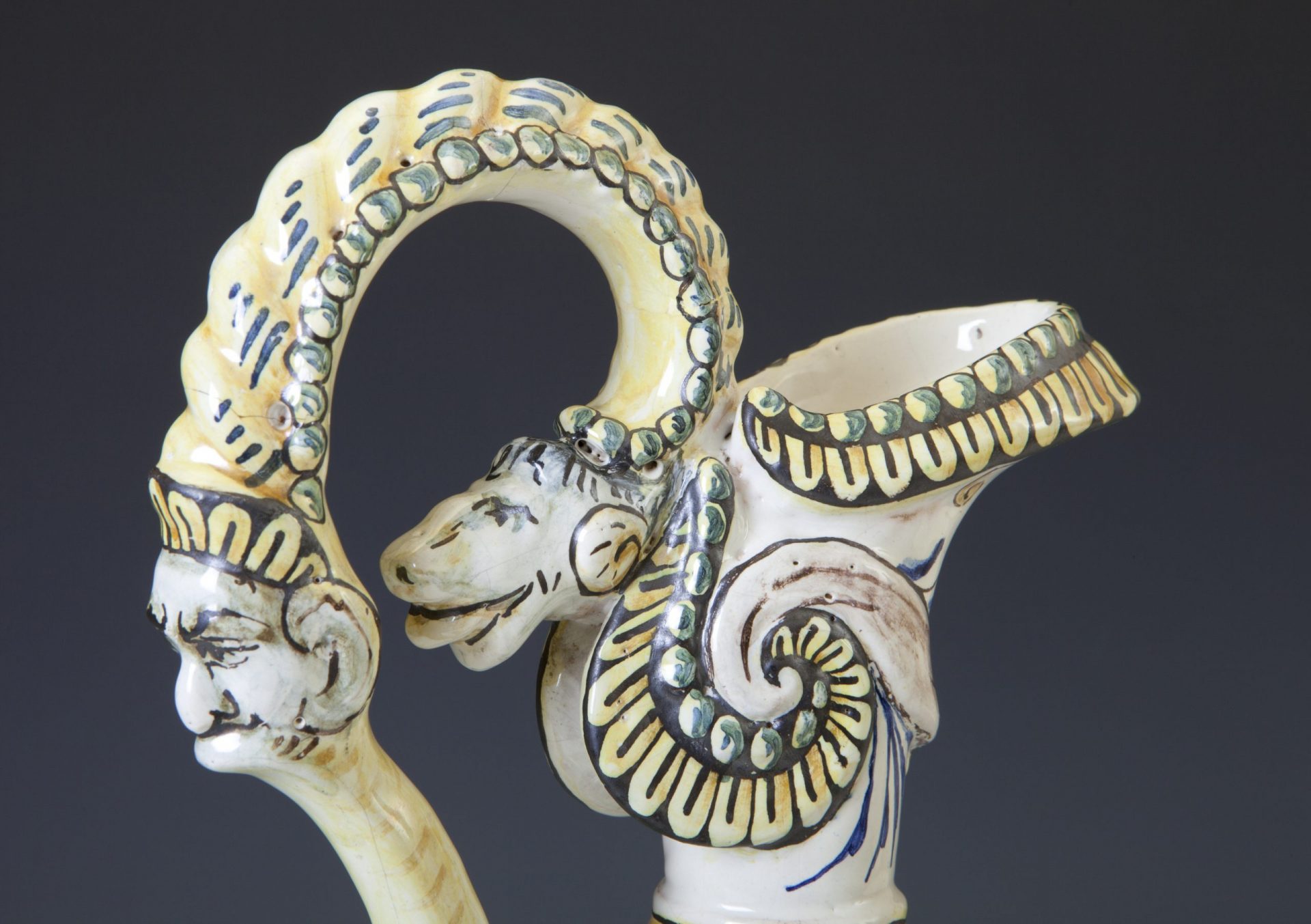Zbliżenie na ozdobny ceramiczny uchwyt i fragment dzbana z stylizowaną twarzą i głową zwierzęcą połączonymi łukiem ozdobionym perełkowymi wypustkami oraz spiralnym motywem muszli i żółto‑zielono‑czarnymi malowanymi wzorami.
