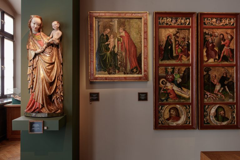 Po lewej wysoka, złocona drewniana rzeźba kobiety w bogato fałdowanej szacie trzymającej nagie dziecko stojąca na zielonym cokole przy oknie; na środku ramowany obraz przedstawiający siedzącą kobietę z dzieckiem i pochyloną postać mężczyzny w czerwonym płaszczu z aureolami i księgą; po prawej dwa pionowe, czerwone panele z wieloma malowanymi scenami, medalionami z popiersiami i złotymi tłami oraz małe tabliczki opisowe na jasnej ścianie.