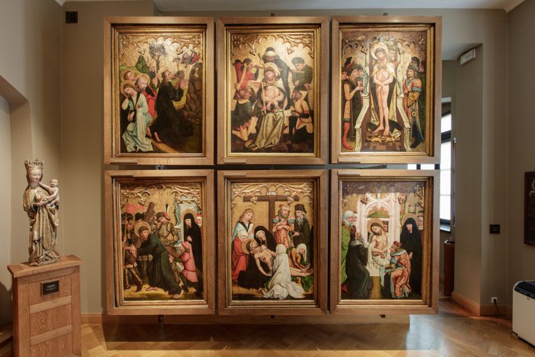 Sześć dużych paneli malarskich w drewnianych ramach ułożonych w dwóch rzędach na ścianie muzealnej, każdy z bogatym złoconym tłem i ornamentyką przedstawiający szczegółowe sceny religijne męki (m.in. koronowanie cierniem, biczowanie, ukrzyżowanie, zdjęcie z krzyża), po lewej stojąca na postumencie drewniana rzeźba koronowanej kobiety trzymającej dziecko z metalową tabliczką, całość na tle beżowych ścian, drewnianej podłogi i okna po prawej.