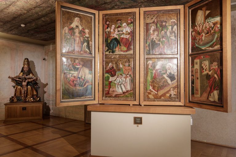 Wnętrze muzeum z rozłożonym, wielodzielnym ołtarzem malowanym na drewnie ukazującym liczne sceny religijne w polach obrazowych, ustawionym na jasnej podstawie na środku pokoju z ozdobnym malowanym stropem i drewnianą podłogą; po lewej stoi polichromowana rzeźba drewniana przedstawiająca siedzącą, brodatą postać z krucyfiksem.