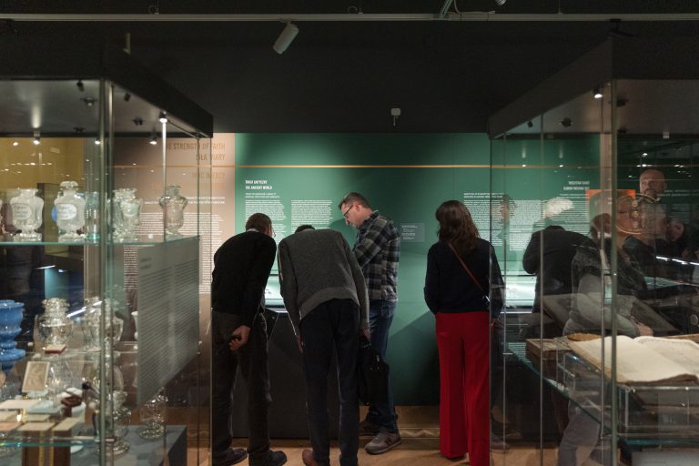 Wnętrze muzeum z ciemnym sufitem i zieloną ścianą wystawową opisaną jako "Świat Antyczny", na środku cztery osoby pochylone nad gablotą, po bokach szklane witryny z aptecznymi naczyniami i innymi eksponatami oraz widoczne odbicia odwiedzających w szybach.