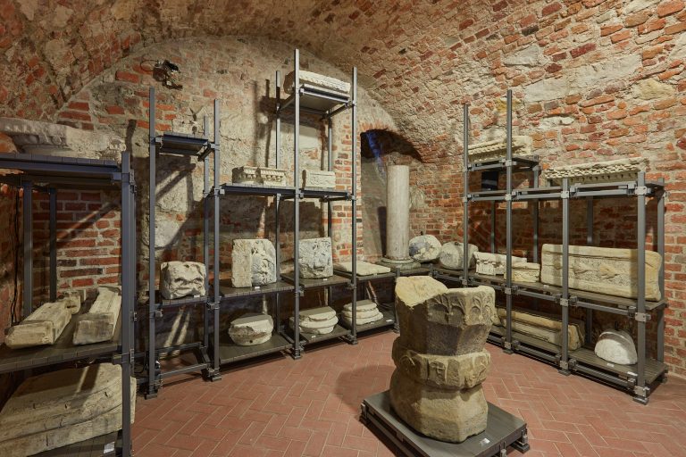 Piwniczne pomieszczenie z ceglanym sklepieniem i terakotową posadzką, w którym metalowe regały i platformy podpierają liczne kamienne fragmenty architektoniczne — kolumny, kapitele, rzeźbione bloki i płaskorzeźby — oraz centralnie stojący masywny, warstwowy blok kamienny.