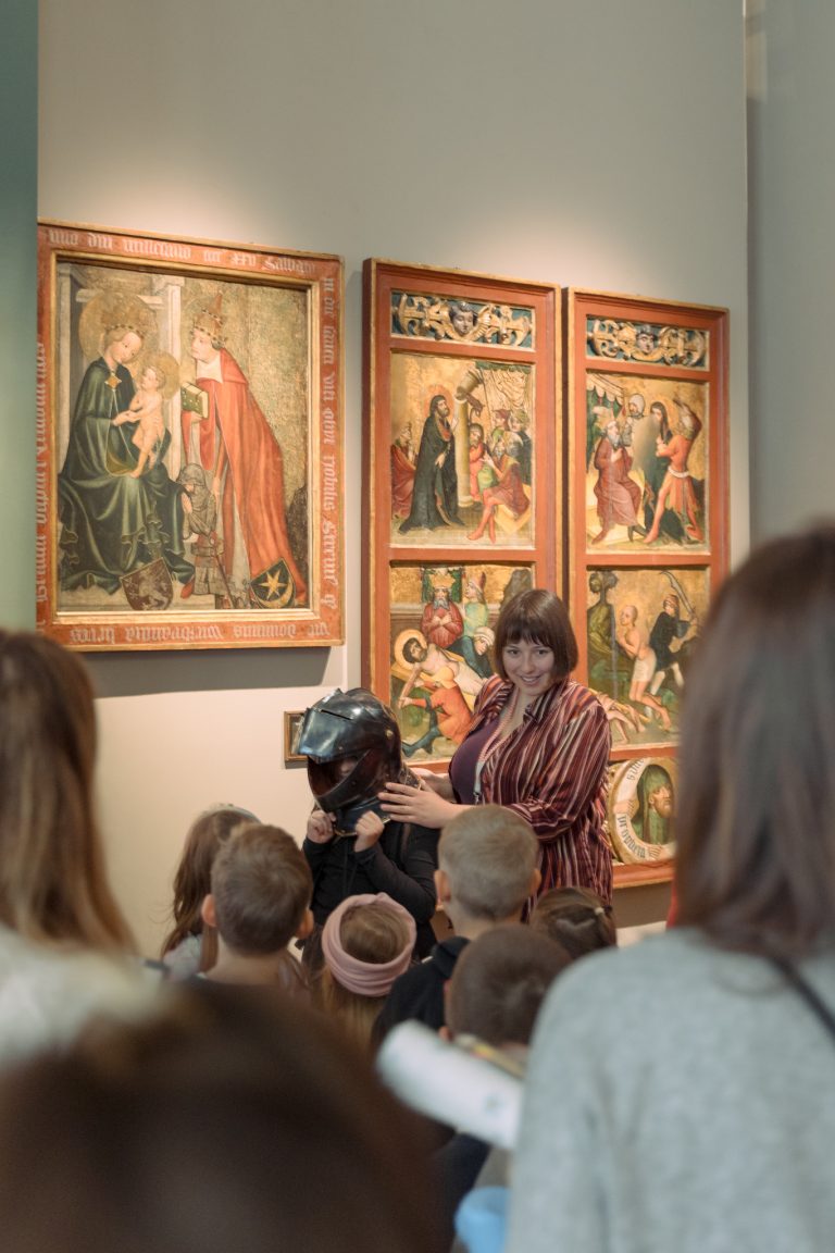 Kobieta pomaga dziecku założyć ciemny metalowy hełm przed zebraną grupą dzieci w muzeum, a w tle na jasnej ścianie wiszą trzy ozdobne drewniane panele z malowanymi scenami religijnymi.