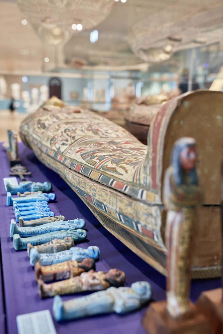 Zbliżenie ozdobnego egipskiego sarkofagu z malowanymi motywami i rzędem małych turkusowych figurek ułożonych na fioletowym materiałem, na rozmytym tle wnętrza muzeum.
