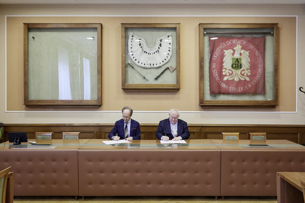 Dwa siedzące przy długim, obitym brązową skórą biurku mężczyźni w garniturach podpisują dokumenty; za nimi na ścianie trzy duże drewniane gabloty — lewa pusta z szybą, środkowa z białym futrzanym kołnierzem i dwoma narzędziami, prawa z czerwonym proporcem z herbem i napisem AGH Akademia Górniczo‑Hutnicza Stanisława Staszica w Krakowie.
