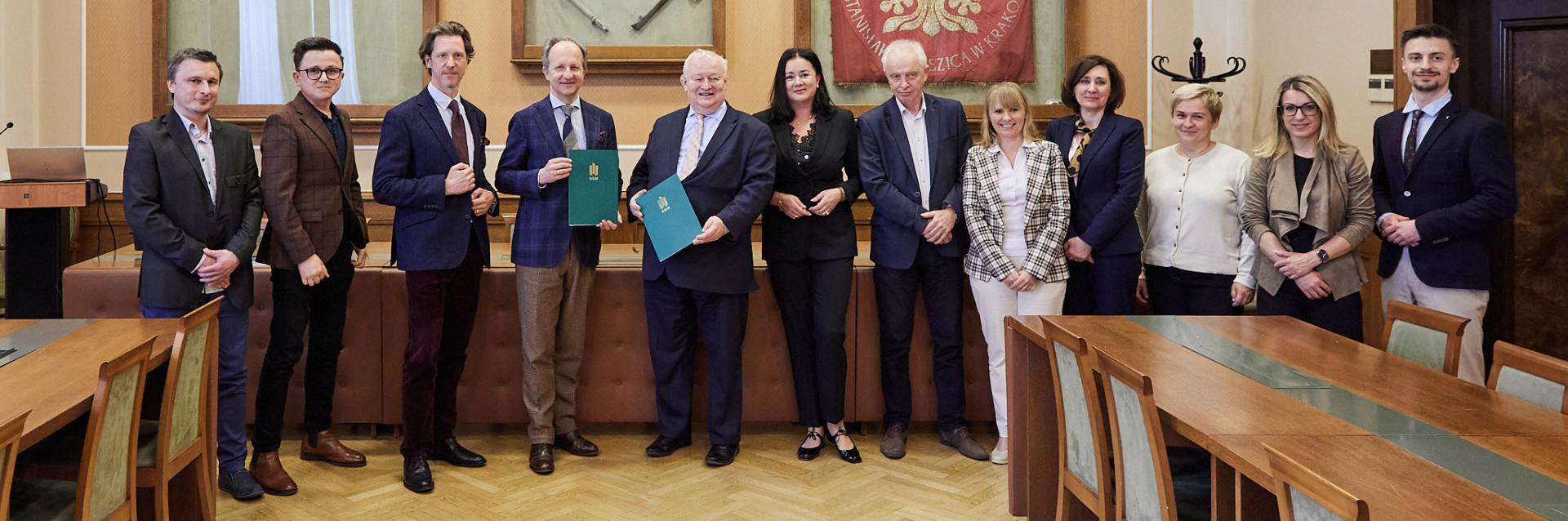 Grupa trzynastu osób w formalnym ubraniu stoi w jednym rzędzie w sali konferencyjnej z drewnianymi stołami i tapicerowanymi krzesłami; na środku dwaj mężczyźni trzymają zielone teczki z złotym emblematem, za nimi wisi czerwony sztandar i oprawiony obraz, po lewej mównica z laptopem, po prawej wieszak na ubrania, całość na jasnym parkiecie.