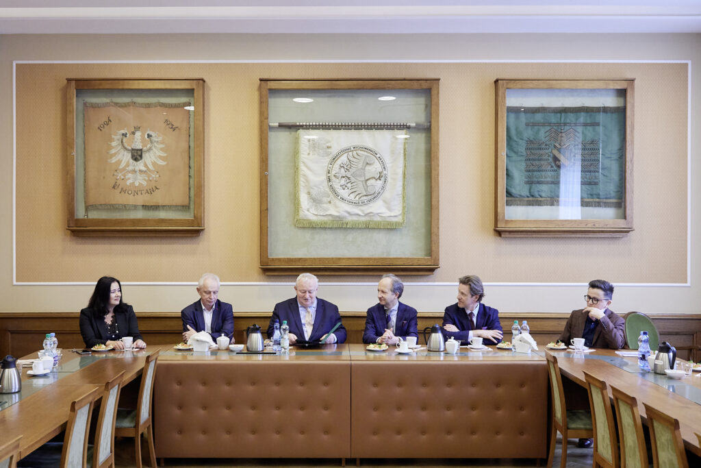 Sześć osób w formalnych ubraniach siedzi przy długim drewnianym stole konferencyjnym z filiżankami, dzbankami, butelkami wody i talerzami z przekąskami; za nimi na beżowej ścianie wiszą trzy oprawione historyczne sztandary w drewnianych ramach, a przednia część stołu ma pikowaną brązową tapicerkę.