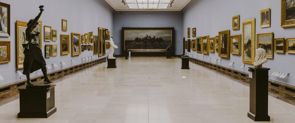 Duża, pusta sala galerii sztuki z jasnym, podłużnym świetlikiem sufitowym, obrazami w złoconych ramach wiszącymi po obu stronach ścian oraz kilkoma rzeźbami na czarnych piedestałach (m.in. ciemnobrązowa postać z uniesioną ręką po lewej, biała marmurowa rzeźba w środku i popiersie po prawej), a na końcu sali duży, ciemny obraz przedstawiający jeźdźców na koniach.