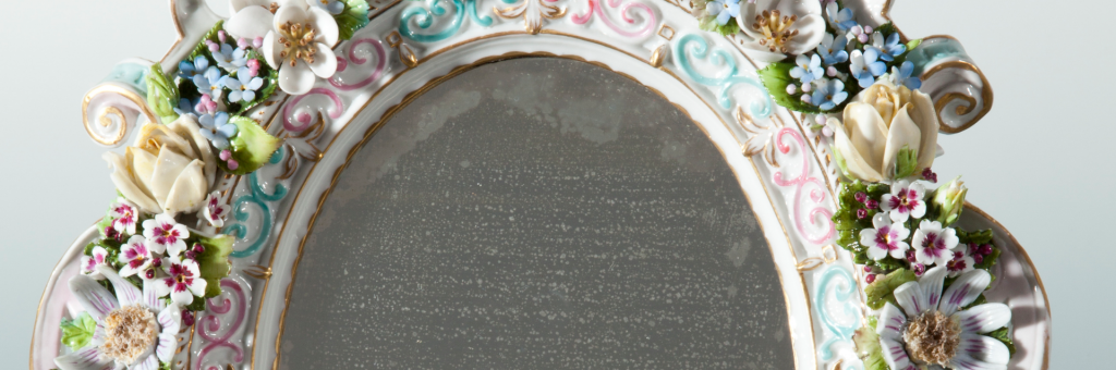 Zbliżenie górnej części owalnej ramy lustra z białej porcelany ozdobionej różowo‑turkusowymi ornamentami i złoceniami, bogato udekorowanej reliefowymi porcelanowymi kwiatami — kremowymi różami, drobnymi niebieskimi i różowymi kwiatuszkami oraz białymi kwiatami przypominającymi stokrotki — z zielonymi listkami i postarzałą, nakraplaną powierzchnią lustra.