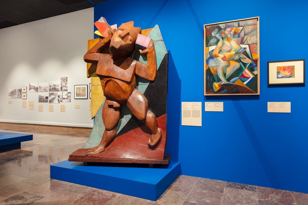 Wnętrze galerii z dużą rzeźbą o geometrycznych, figuratywnych kształtach wykonanej z drewna w rdzawym odcieniu ustawioną na niebieskim podeście na pierwszym planie, za nią wielobarwna, geometryczna tło-płyta; po prawej na niebieskiej ścianie dwa oprawione, kolorowe obrazy o kubistycznej stylistyce z tabliczkami informacyjnymi, po lewej biała ściana z czarno-białymi fotografiami i opisami oraz marmurowa posadzka.
