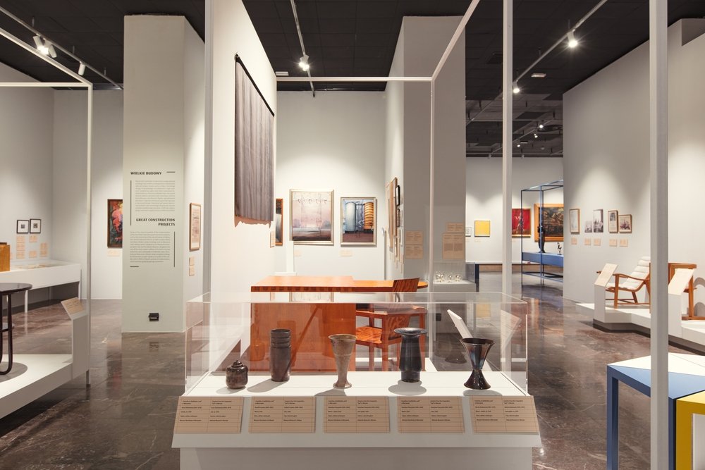 Wnętrze jasnej galerii wystawowej z białymi ścianami i czarnym sufitem, na pierwszym planie oszklona gablotka z kilkoma ceramicznymi naczyniami i opisami, za nią drewniany stół i krzesła oraz obrazy i teksty ekspozycyjne na ścianach, a w głębi kolejne wnęki z meblami i rzeźbami na błyszczącej ciemnej posadzce.