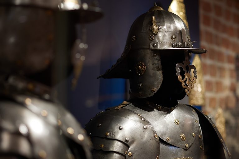 Zbliżenie na stalowy hełm i napierśnik zbroi ozdobione złotymi nitami i metalowymi ornamentami; hełm ma daszek i ażurową złotą przyłbicę, w tle rozmyta ceglana ściana i fragment innego pancerza.