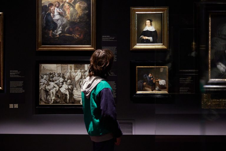 Osoba widziana od tyłu w zielonej kamizelce i z kapturem stoi przed ciemną ścianą galerii, na której w złotych ramach wiszą cztery obrazy — u góry po lewej scena religijna, u góry po prawej portret kobiety w białym kołnierzu, poniżej po lewej monochromatyczna scena tłumu, a po prawej mała scena przy stole, oraz w prawym panelu widać odbicie czyjejś twarzy w szybie.