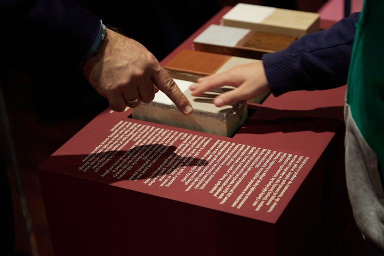 Większa dłoń z obrączką i opaską na nadgarstku wskazująca palcem na prostokątną teksturowaną próbkę materiału, obok mniejsza dłoń uniesiona nad szeregiem prostokątnych próbek w odcieniach beżu, brązu i ceglanej czerwieni ustawionych na nachylonym czerwonym pulpicie z białym tekstem.