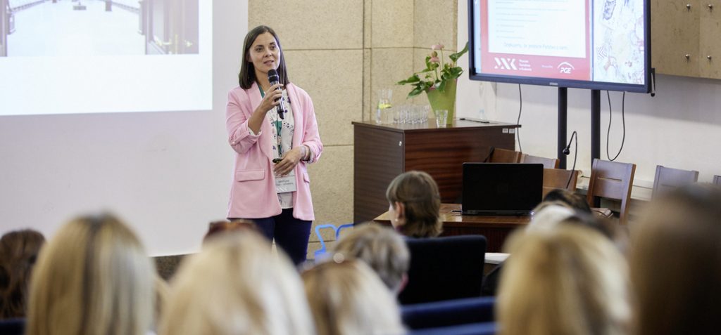 Kobieta w różowej marynarce stoi z mikrofonem przed ekranem projekcyjnym po lewej i dużym monitorem po prawej w sali konferencyjnej, obok stolik z laptopem, wazonem z kwiatami i szklankami, a w pierwszym planie siedzący uczestnicy.
