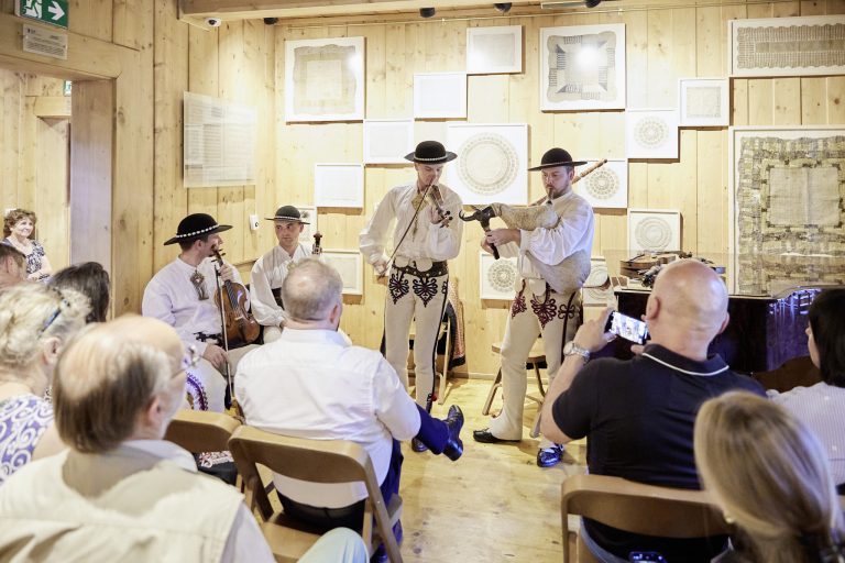 W małej sali z jasnymi drewnianymi panelami i oprawionymi tkaninami na ścianach grupa muzyków w haftowanych, jasnych strojach i czarnych kapeluszach gra — dwóch na skrzypcach i jeden na dudach — przed nimi siedzi publiczność, a jedna osoba robi zdjęcie telefonem.