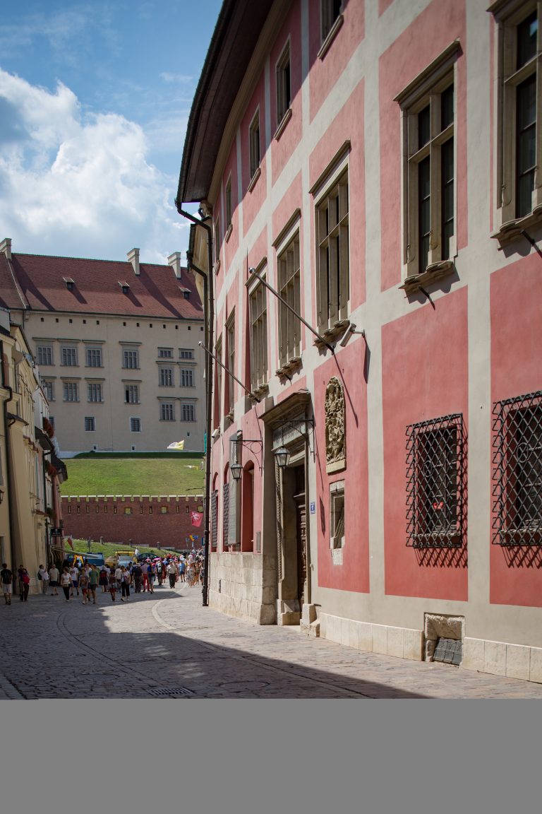 Uliczka z brukowaną nawierzchnią, po prawej duża różowo‑biała kamienica z wysokimi oknami, metalowymi kratami i kamiennym portalem, po lewej w oddali czerwony mur warowny i zielony stok prowadzący do dużego jasnego budynku, grupa pieszych oraz niebieskie niebo z chmurami.