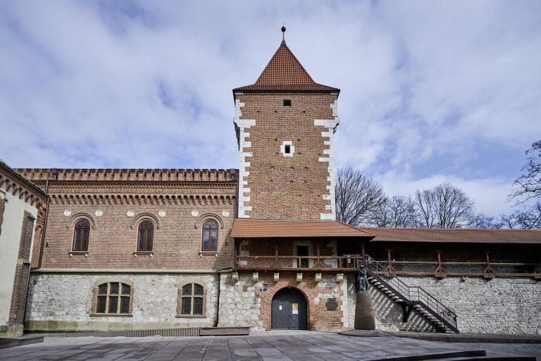 Widok fasady murowanego budynku z wysoką ceglano‑kamienną wieżą nakrytą czerwonym dachem z dachówek, ozdobnymi łukowymi oknami i detalami z białego kamienia na narożnikach, kamienną dolną częścią z małymi oknami, drewnianym balkonem nad wejściem, zewnętrznymi metalowymi schodami po prawej, stojącym przy nich pomnikiem oraz wybrukowanym dziedzińcem na pierwszym planie, wszystko na tle nieba i nagich drzew.