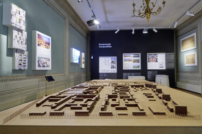Widok wnętrza wystawy z dużą drewnianą makietą urbanistyczną na pierwszym planie, w głębi czarna ściana z napisem „Kompozycja / Composition”, po bokach podświetlane panele z fotografiami, ozdobne listwy ścienne, sufit z żyrandolem i reflektorami ekspozycyjnymi.