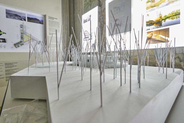 Model architektoniczny na białej platformie przedstawiający miniaturowy budynek z tarasem otoczony gęstą siatką cienkich pionowych prętów przypominających drzewa, a w tle jasne plansze z planami i zdjęciami.