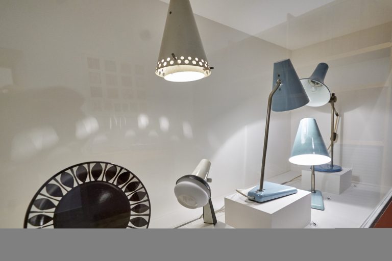 Ekspozycja lamp na białym tle: centralnie wiszący biały stożkowy klosz z perforowanym brzegiem, po prawej kilka niebieskich lamp biurkowych i podłogowych o stożkowych kloszach ustawionych na białych piedestałach, z przodu mała biała lampka na regulowanym ramieniu i po lewej fragment okrągłego czarnego siedziska z dekoracyjnym, wyciętym wzorem.