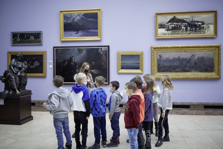 Grupa dzieci w maseczkach stoi w galerii przed jasnofioletową ścianą ozdobioną kilkoma oprawionymi obrazami, po lewej duża ciemna rzeźba, a wśród dzieci stoi młoda osoba w maseczce.