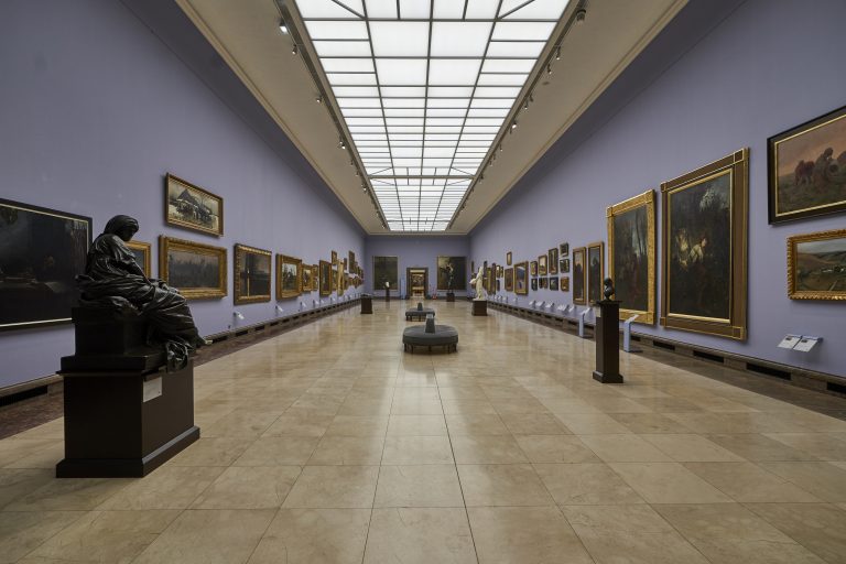 Długa, symetryczna sala galerii z dużym prostokątnym świetlikiem w suficie, jasną marmurową posadzką, fioletowymi ścianami obwieszonymi wieloma obrazami w ozdobnych ramach oraz kilkoma rzeźbami na postumentach i okrągłymi siedziskami ustawionymi w środkowej osi.