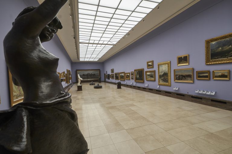 Widok długiej sali galerii z dużym prostokątnym świetlikiem sufitowym, fioletowymi ścianami obwieszonymi obrazami w pozłacanych ramach, zbliżenie ciemnej brązowej rzeźby po lewej, biała rzeźba na postumencie w głębi, jasna kamienna posadzka i kilka ławek pośrodku.