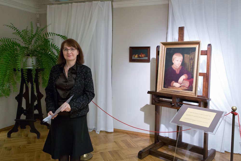 Kobieta w ciemnej kurtce i spódnicy trzymająca papiery stoi po lewej stronie na drewnianej podłodze przed białymi zasłonami; po jej lewej duża paproć w białej doniczce na ozdobnym ciemnym stojaku, a po prawej na drewnianym sztalugu oprawiony portret dziecka, obok tablica informacyjna, czerwona lina odgradzająca i mały obrazek na ścianie.